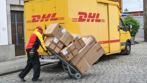 Ein DHL-Bote bringt einen Stapel Pakete per Sackkarre zum Lieferfahrzeug. 