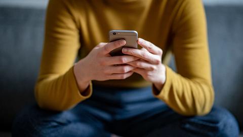 Eine Frau befasst sich mit ihrem Handy auf einer Couch sitzend.