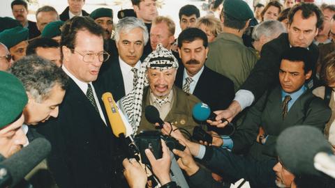 Der frühere Außenminister Klaus kinkel und Palästinenserführer Jassir Arafat umringt von einem Pulk Journalist*innen mit mikrofonen, darunter auch Christopher Plasss mit hr-Mikrofon, Aufnahme von 1997