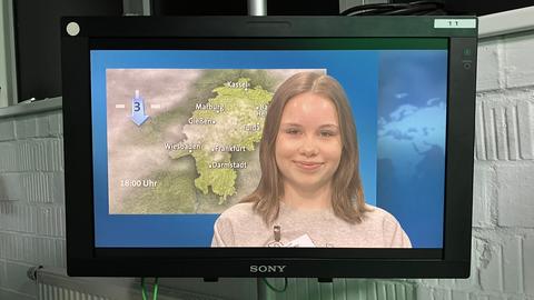 Fernsehmonitor vor einer Wand, darauf ist ein Mädchen vor einem Wetterkarten-Hintergrund zu sehen