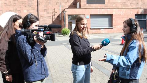 Im Außengelände: Ein Mädchen mit VJ-Kamera auf der Schulter und ein Mädchen mit einem Handmikrofon interviewen ein anderes Mädchen, das ein Tongerät umgehängt hat und Kopfhöärer trägt. Faneben helfend eine hr-Mitarbeiterin.